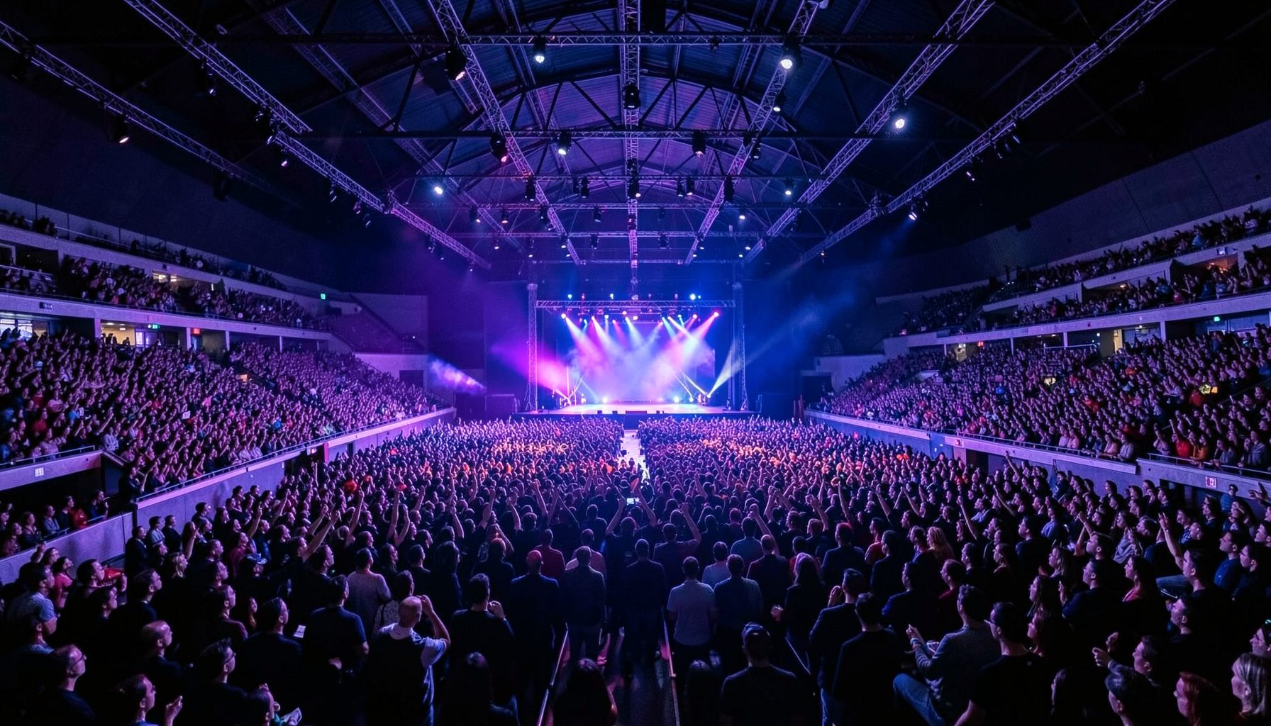 A wide interior view of a large modern arena with a stage at one end, dramatic purple and blue stage lighting sweeping across the crowd